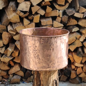 Könnte beinhalten: Ein gehämmerter Kupfertopf steht auf einem Baumstumpf vor einem Hintergrund aus gestapeltem Brennholz.