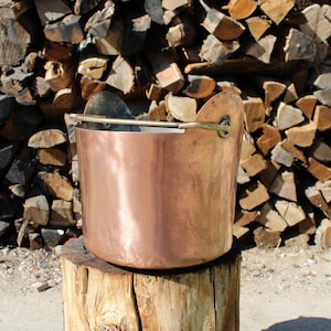 Puede incluir: Una olla de cobre con un asa de latón, sentada sobre un tocón de árbol frente a una pila de leña.