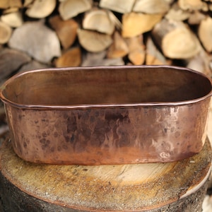 May include: A hammered copper planter with an oval shape. The planter is a warm copper color and sits on a tree stump. The background features a stack of firewood, suggesting an outdoor or rustic setting.