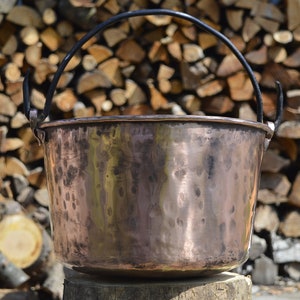 May include: A large copper pot with a black metal handle. The pot is sitting on a wooden stump in front of a pile of firewood.