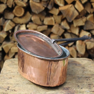 Pode incluir: Uma panela oval de cobre vintage com tampa e alça. A panela está sobre uma superfície de madeira com uma pilha de lenha no fundo.
