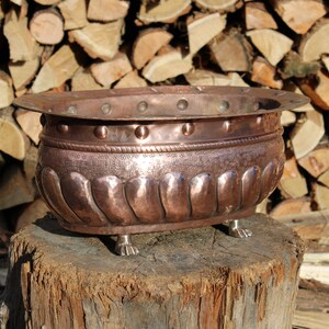 May include: A copper planter with a hammered finish and decorative details. It has a wide, oval shape and sits on four feet. The planter is on a wooden stump.