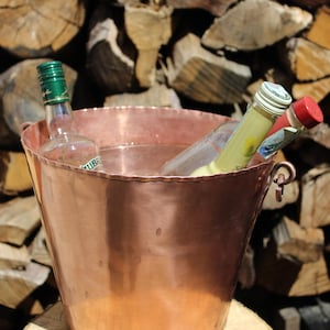Peut inclure: Un seau à glace en cuivre rempli d'eau et de plusieurs bouteilles de boissons. Le seau a une finition brillante et est placé devant un arrière-plan de bois de chauffage empilé. Les bouteilles ont des bouchons et des étiquettes de couleurs variées.