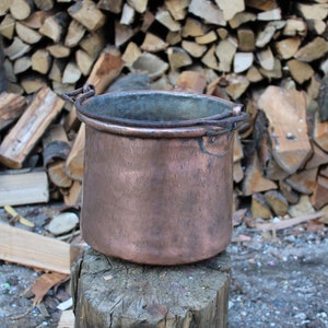 May include: A copper pot with two iron handles sits on a wooden stump. The pot is round and has a slightly flared lip. The stump is weathered and has a rough texture.