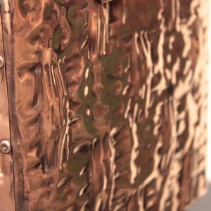 May include: A rectangular copper object with a highly reflective, hammered, and undulating surface texture. The side panel is attached with two visible silver-toned metal screws, creating a distinct contrast against the warm, metallic copper finish.