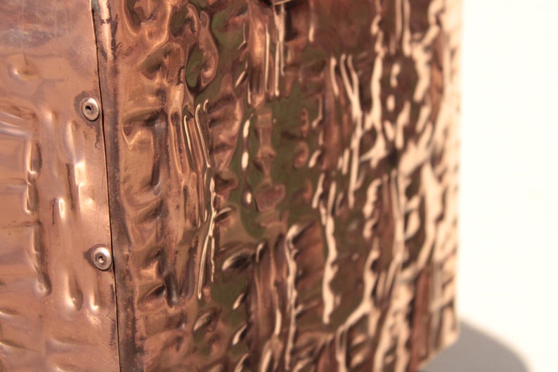 May include: A rectangular copper object with a highly reflective, hammered, and undulating surface texture. The side panel is attached with two visible silver-toned metal screws, creating a distinct contrast against the warm, metallic copper finish.