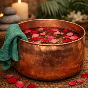 May include: A large, round, hammered copper basin filled with water and scattered pink and red rose petals. A teal-colored towel is draped over the rim of the basin. The copper surface has a textured, metallic finish with warm, reflective tones.