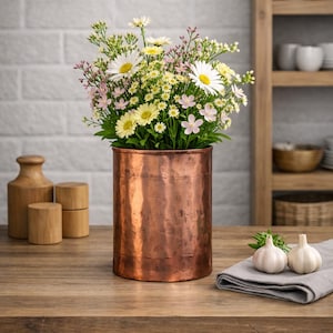 Könnte beinhalten: Eine zylindrische Vase aus gehämmertem Kupfer, gefüllt mit einem Strauß aus weißen Margeriten, gelben Blumen und kleinen rosa Blüten. Die Vase hat eine metallische Oberfläche mit sichtbarer Textur und zeigt ein lebendiges Arrangement aus frischen Pflanzen.