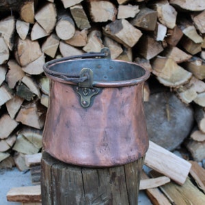 Puede incluir: Una olla de cobre con un asa de latón y una base de madera. La olla está sentada sobre un tronco de árbol frente a una pila de leña.