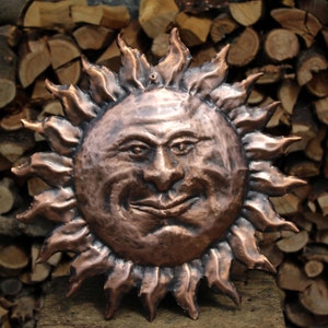 May include: A copper sun sculpture with a face and a textured surface. The sun is surrounded by rays.