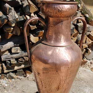 Könnte beinhalten: Eine große Kupfervase mit zwei Henkeln. Die Vase hat eine gehämmerte Textur und ist in einem satten Kupferton gehalten.