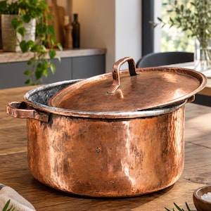 May include: A large, round, hammered copper pot with a matching lid and two side handles. The exterior has a textured, weathered copper finish, while the interior shows a metallic silver-toned lining. The lid features a central handle.