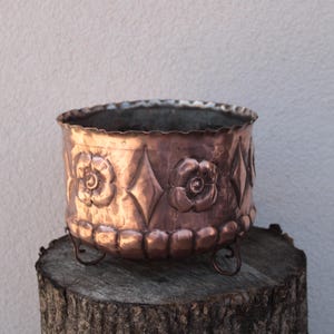 May include: A round copper planter featuring a scalloped top edge and a decorative embossed pattern of alternating flowers and diamonds. The base of the vessel is finished with a row of rounded indentations and stands on three small metal feet.