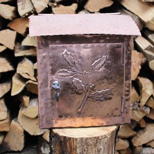 May include: A copper mailbox with a holly leaf design. The mailbox has a hinged door and a small keyhole. It is sitting on a wooden stump.