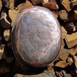 May include: A large, antique copper cooking pot with a hammered finish. The pot has a rounded shape and a dark, mottled patina. It rests on a weathered wooden stump, with a backdrop of stacked firewood. The copper pot is a rustic kitchenware item.