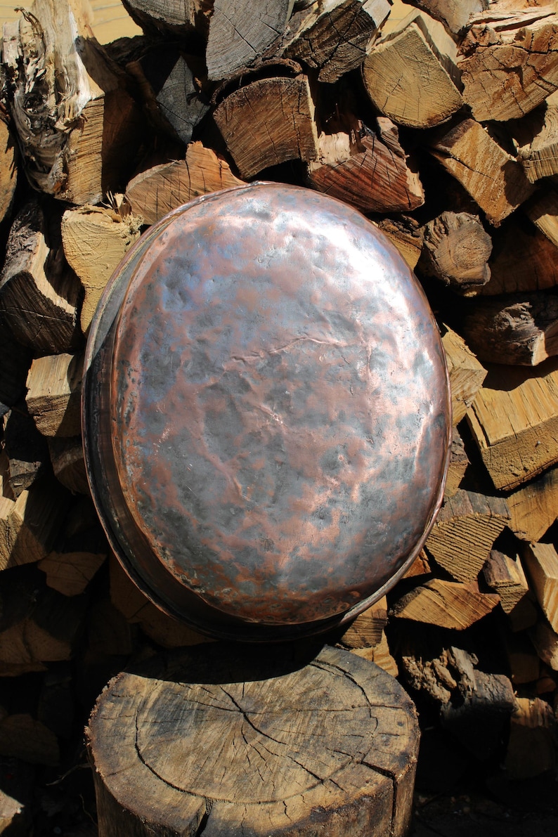 May include: A large, antique copper cooking pot with a hammered finish. The pot has a rounded shape and a dark, mottled patina. It rests on a weathered wooden stump, with a backdrop of stacked firewood. The copper pot is a rustic kitchenware item.