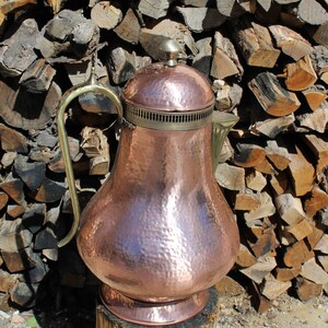 Op de afbeelding: Een gehamerde koperen theepot met een messing deksel en handvat. De theepot staat op een boomstronk.