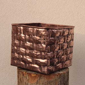 May include: A copper-toned, rectangular basket with a woven pattern. The basket sits on a wooden stump, highlighting its handcrafted nature. The design suggests it could be used for storage or as a decorative planter, with dimensions of approximately 20 cm.