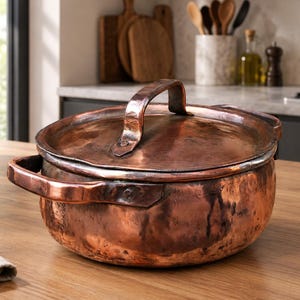 Puede incluir: Una olla redonda de cobre martillado con tapa a juego y dos asas laterales. La superficie metálica presenta un acabado marrón cobrizo cálido con una textura visible y marcas que denotan un proceso de fabricación artesanal.
