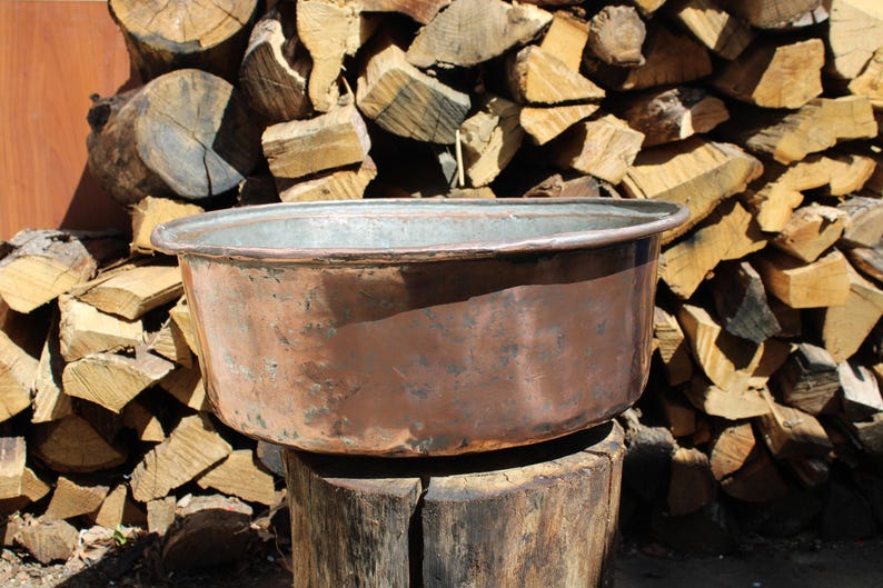 May include: An aged copper basin with a wide rim, displaying signs of wear. The basin is placed on a weathered wooden base, with a backdrop of stacked firewood. The copper has a rich, reddish-brown patina, showing its age.