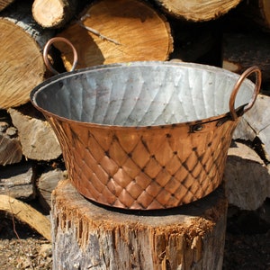 May include: A large copper pot with two handles sits on a tree stump. The pot has a hammered finish and a diamond pattern on the bottom.