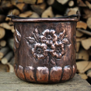 Può includere: Un secchio di rame con finitura martellata e design floreale. Il secchio ha un manico ed è appoggiato su una superficie di legno.