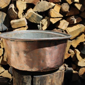 May include: A copper basin with a wide rim, showing signs of weathering, set against a backdrop of stacked firewood. The basin's surface has variations in colour, from reddish-brown to darker hues. The rustic setting suggests outdoor use or a vintage aesthetic.