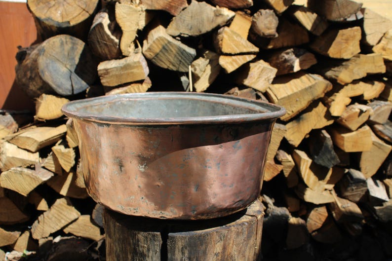 May include: A copper basin with a wide rim, showing signs of weathering, set against a backdrop of stacked firewood. The basin's surface has variations in colour, from reddish-brown to darker hues. The rustic setting suggests outdoor use or a vintage aesthetic.