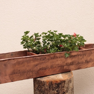 May include: A rectangular copper planter box with three compartments, filled with green plants and red flowers. The planter is placed on a tree stump against a neutral wall. The copper has a weathered, aged appearance.
