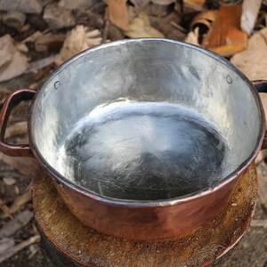 May include: A copper pot with two handles, the inside is lined with a shiny silver material. The pot is sitting on a wooden stump.