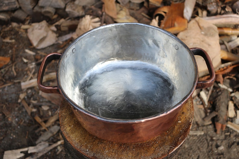 May include: A copper pot with two handles, the inside is lined with a shiny silver material. The pot is sitting on a wooden stump.