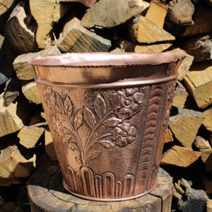 May include: A copper-colored metal planter with embossed floral and leaf designs. The planter has a textured surface and a decorative rim. The background features a stack of firewood.