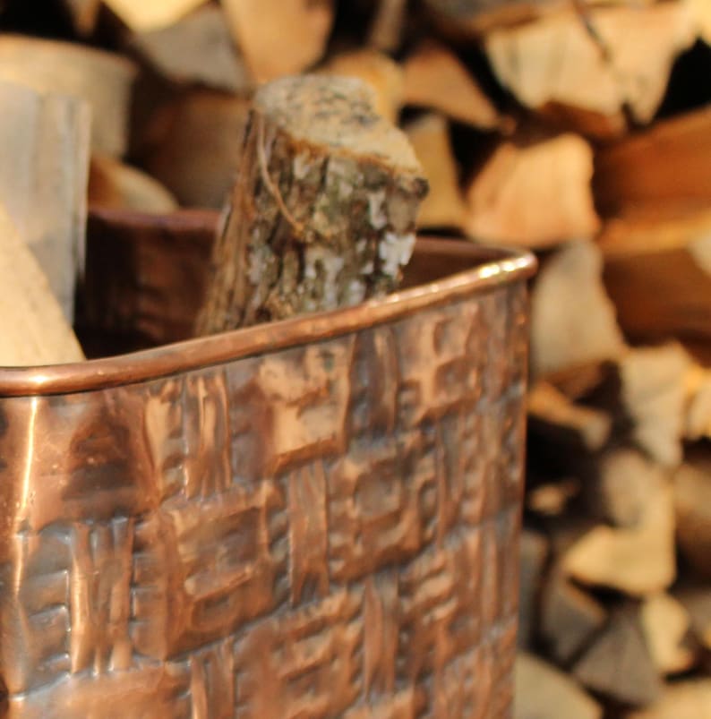 May include: A rectangular copper container featuring a textured, embossed pattern on its exterior surface. The metallic vessel has a smooth, rounded rim and is filled with pieces of natural wood logs, showcasing a warm, reflective finish.
