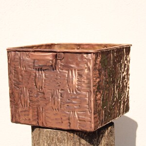 May include: A square-shaped copper storage bin featuring a textured, embossed basket-weave pattern on its exterior surfaces. The container includes a small, flat handle attached to the upper side and exhibits a warm, metallic reddish-brown finish.