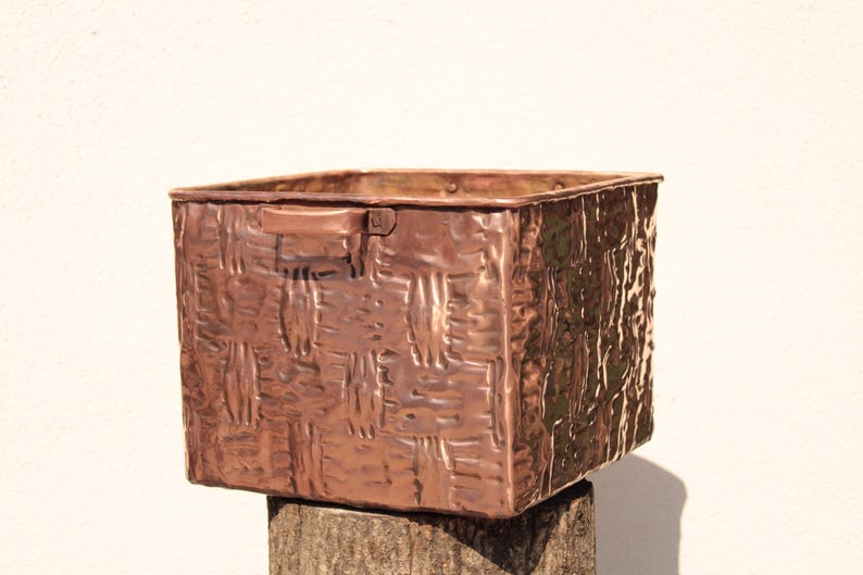 May include: A square-shaped copper storage bin featuring a textured, embossed basket-weave pattern on its exterior surfaces. The container includes a small, flat handle attached to the upper side and exhibits a warm, metallic reddish-brown finish.