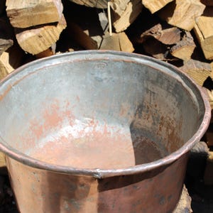 May include: A large, round, open-top copper basin with a wide, rolled rim. The exterior features a warm, metallic copper finish, while the interior shows a weathered, silvery-grey patina with visible signs of oxidation and wear throughout.