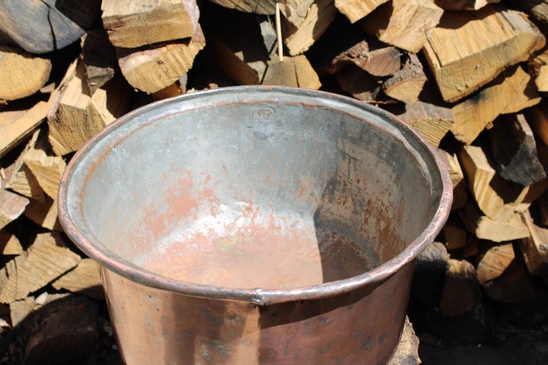 May include: A large, round, open-top copper basin with a wide, rolled rim. The exterior features a warm, metallic copper finish, while the interior shows a weathered, silvery-grey patina with visible signs of oxidation and wear throughout.