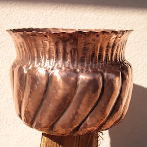 Könnte beinhalten: Handgefertigter Kupfertopf mit strukturierter, gehammerter Oberfläche. Der Topf hat einen breiten, wellenförmigen Rand und ein spiralförmiges, geripptes Design am Körper. Das Kupfer hat einen warmen, rötlich-braunen Farbton und steht auf einem Holzsockel.
