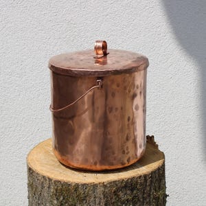 May include: A copper bucket with a lid and a handle. The bucket is cylindrical and sits on a tree stump. The copper has a hammered texture and a warm, reddish-brown colour. The lid has a small loop handle.