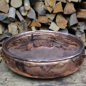 May include: A large, round copper pot with a lid and two handles. The pot is sitting on a wooden surface in front of a pile of firewood.