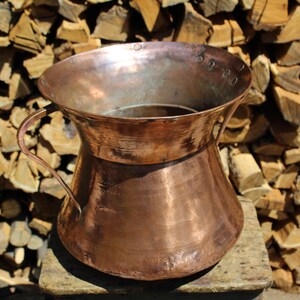 May include: A large copper pitcher with two handles. The pitcher has a wide mouth and a rounded bottom. It is sitting on a wooden surface.
