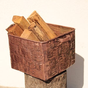 May include: A square copper bin with a textured, woven-look exterior and two side handles. The container is filled with several pieces of light brown, rough-cut firewood logs. The metal surface has a warm, metallic reddish-brown finish.