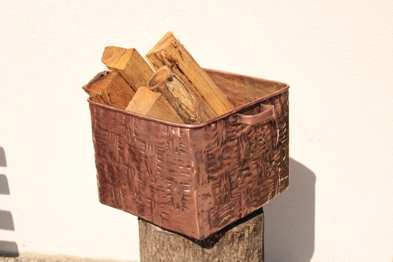 May include: A square copper bin with a textured, woven-look exterior and two side handles. The container is filled with several pieces of light brown, rough-cut firewood logs. The metal surface has a warm, metallic reddish-brown finish.