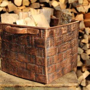May include: A square copper basket with a textured, woven-look exterior pattern and two side handles. The metal surface features a dark, weathered patina with visible indentations and a deep reddish-brown metallic finish, filled with cut firewood.