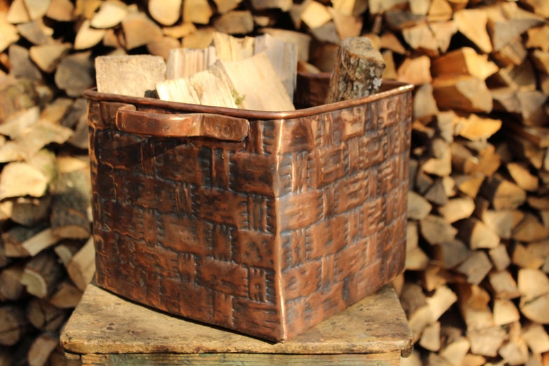 May include: A square copper basket with a textured, woven-look exterior pattern and two side handles. The metal surface features a dark, weathered patina with visible indentations and a deep reddish-brown metallic finish, filled with cut firewood.