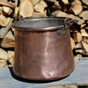 Peut inclure: Un chaudron en cuivre avec une anse, sur fond de bois de chauffage empilé. Le pot a un corps arrondi et une patine sombre et vieillie. La poignée est fixée avec des rivets.