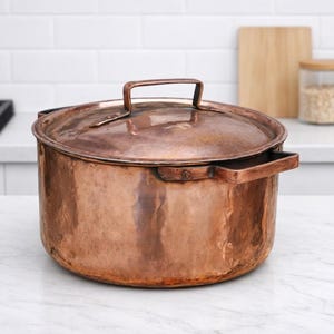May include: A round, hammered copper pot with a matching lid and two side handles. The pot features a warm, metallic finish with visible texture from the hammering process. The lid includes a central rectangular handle for easy lifting.