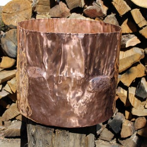 May include: A hammered copper planter with a cylindrical shape. The copper has a rich, reddish-brown color and a textured surface. The planter is set against a backdrop of stacked firewood, suggesting an outdoor or rustic setting.
