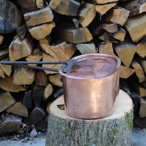 Puede incluir: Una olla de cobre con un mango largo, apoyada sobre un tocón de árbol. La olla tiene una textura martillada y una tapa. Detrás, una pila de leña crea un ambiente rústico. El color de la olla es marrón rojizo cálido.