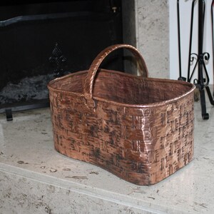 May include: A copper log basket with a woven design. The basket has a handle and is sitting on a light gray surface.